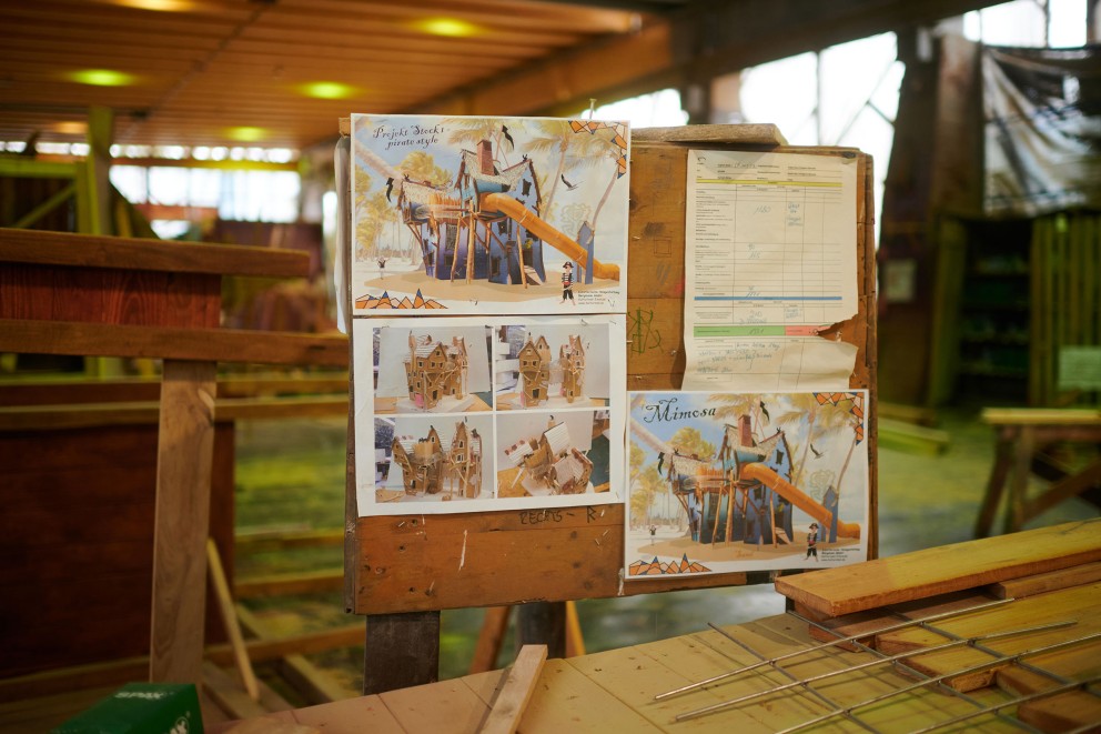 
			Auf einer Tafel in der Werkstatt hängen Skizzen eines Spielhauses mit Rutsche.

		