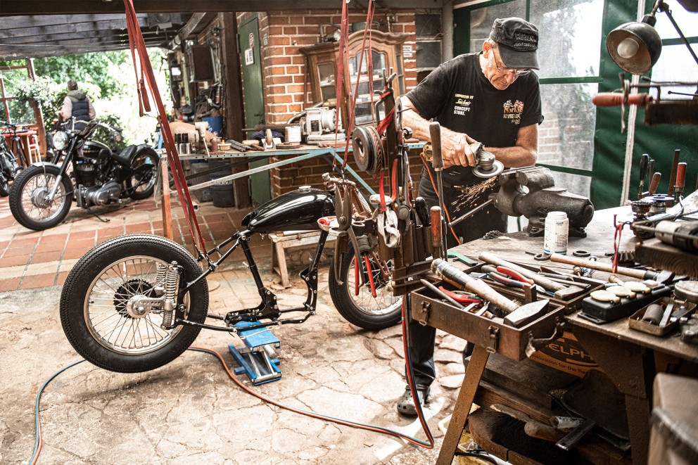 
				Mann flext ein Stück Stahl in der Werkstatt. Im Hintergrund zwei Motorräder.

			